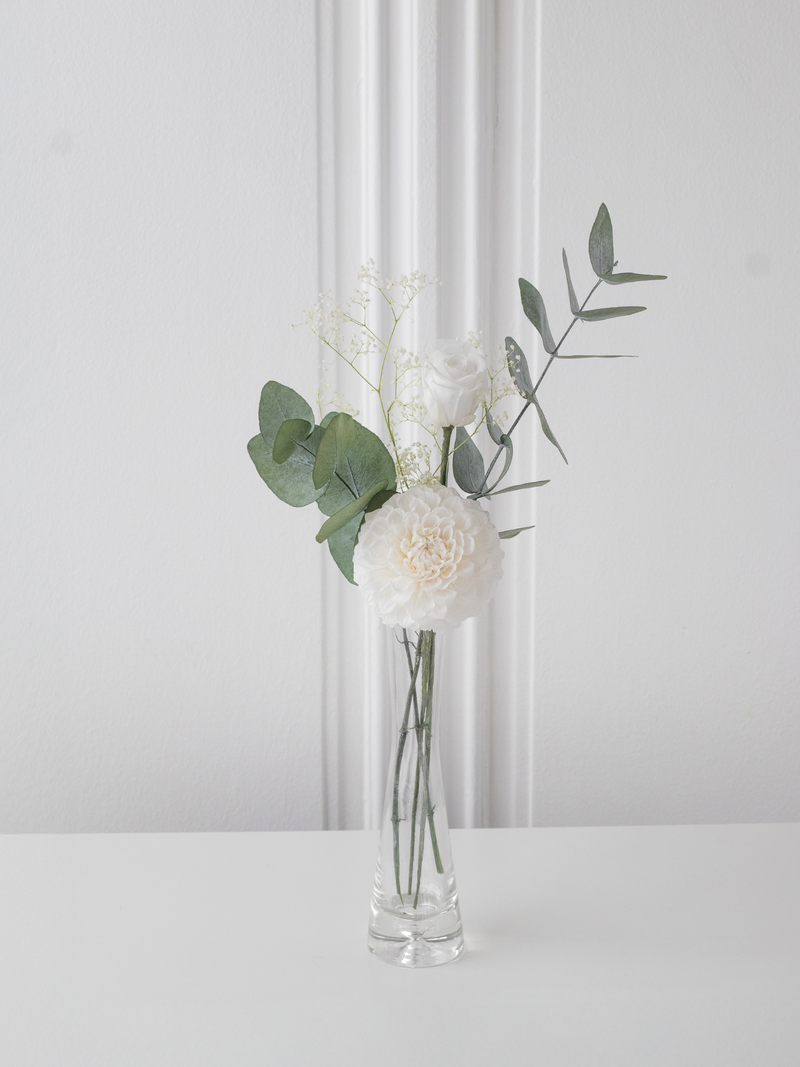Clear glass vase with white preserved flowers and green leaves on a white surface and background. By STILLA for M/Y Vertige.