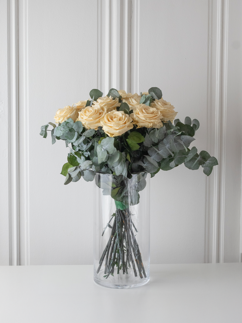 Bouquet of cream preserved roses and eucalyptus in a clear vase against a white wall. By STILLA for M/Y Vertige.